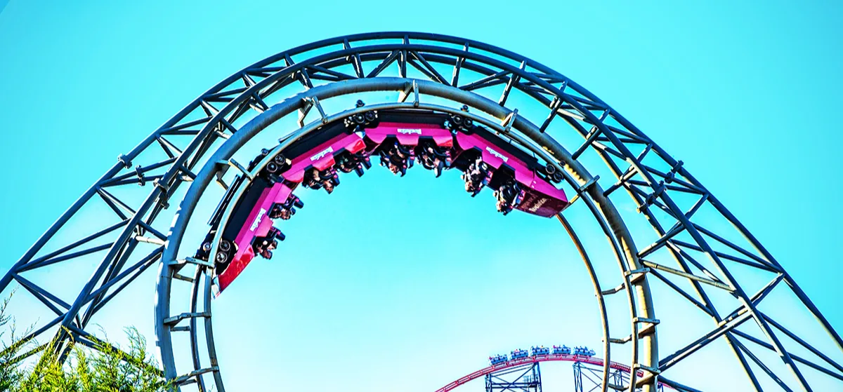 Roller coasters at a theme park with passengers suspended upside down in a bright pink carriage on a loop. 