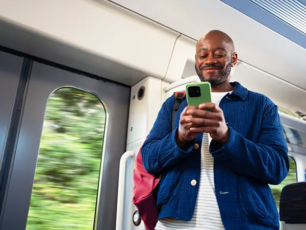 A smiling black man with a bald head and beard, standing on a train and looking at his mobile phone. 