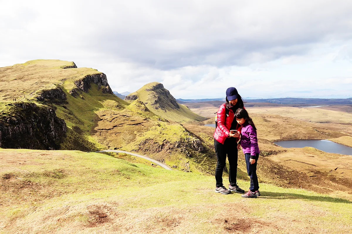 An East Asian mother and young daughter dressed in walking clothes and consulting a phone while standing on a deserted hillside looking over rugged scenery. 