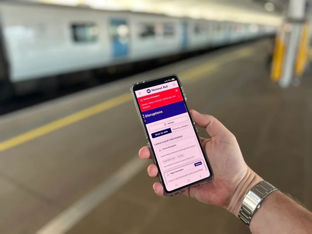 A hand holding a smartphone displaying the National Rail app's Disruptions page, on a train platform.