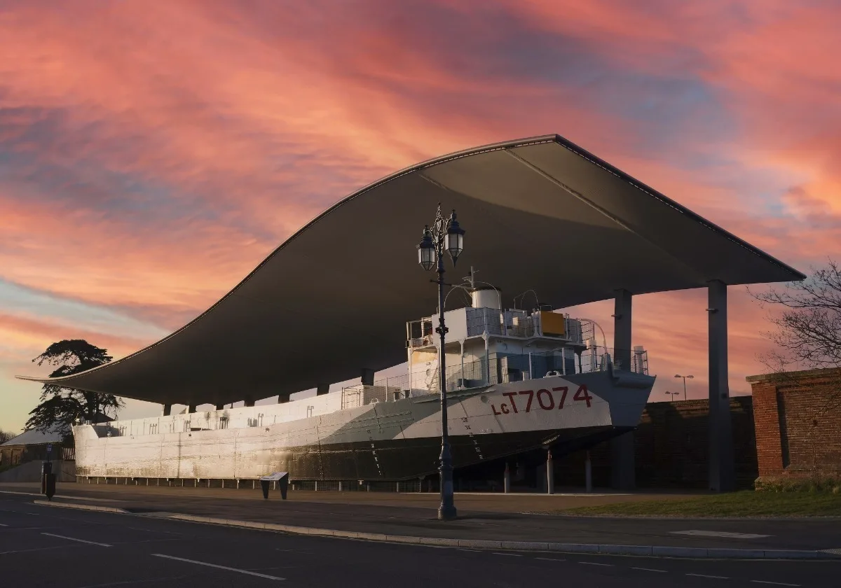 A military landing craft underneath a large modern canopy roof, against a pink sunset. 