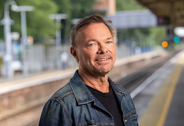 Pete Tong, a middle-aged man in a denim jacket smiling at a railway station platform, with tracks and signal lights visible behind him.