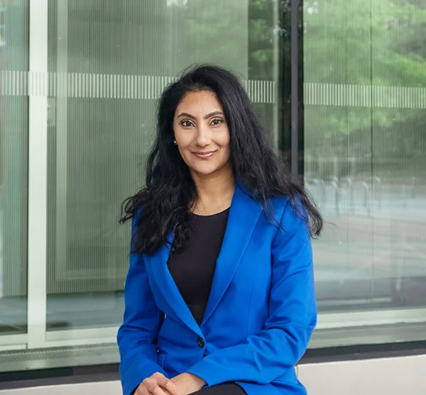 A South Asian woman with long black hair, in a bright blue blazer and black top, smiling in front of glass windows.