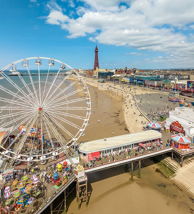 Aerial view of Blackpool beach on a sunny day, with a ferris wheel on a pier visible in the foreground and the seafront and Blackpool Tower in the background.