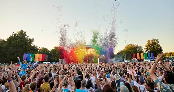 A crowd looking at a stage at an open air concert in daylight, rainbow flags are hanging on either side and rainbow glitter is exploding into the sky.