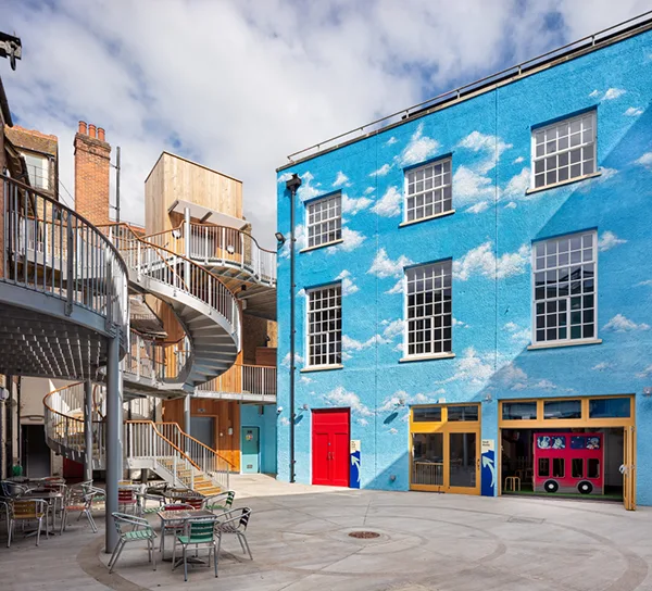 A square with a large steel spiral staircase on one side and a 3-storey building painted with blue sky and white clouds on the other.