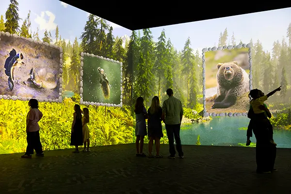 Visitors silhouetted against large immersive wall projections of wildlife, including a bear and forest scenes, in a darkened exhibition space.