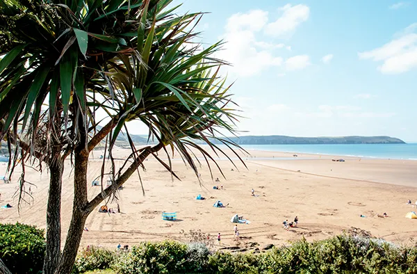 View over a sandy beach dotted with people, with bright blue sea to the right and green hills in the distance, on a sunny day.