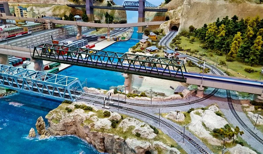 A detailed model railway layout featuring bridges, blue waterways, miniature trains, tunnels, rocky coastlines, and tiny buildings with green scenery.