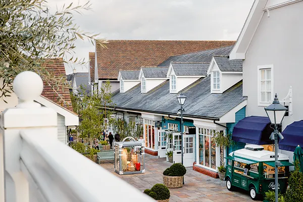 View of an open-air pedestrianised shopping street with 2-storey shops with white fronts and grey roofs. 