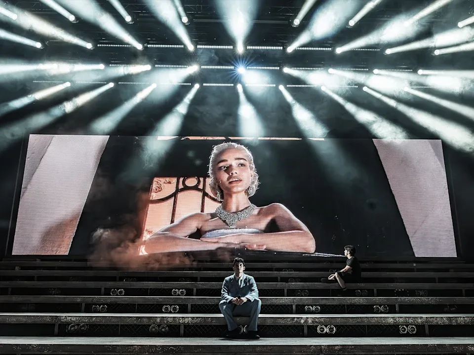 Actress Rachel Ziegler projected on a screen on stage, with 2 actors sitting on steps in front. 