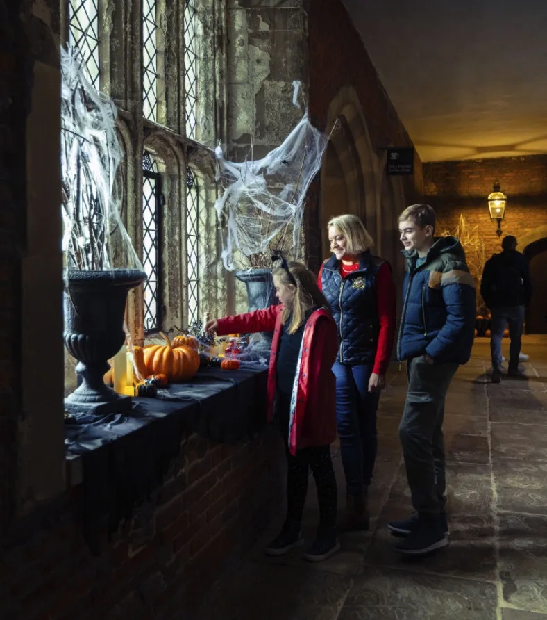 A family exploring Halloween decorations including pumpkins and cobwebs by a leaded window in a dimly lit historic building.