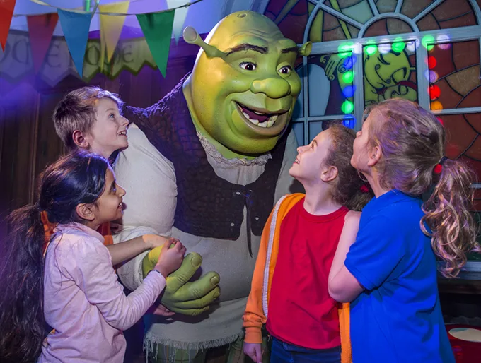 A group of smiling children surround someone in a Shrek costume (a green ogre). 