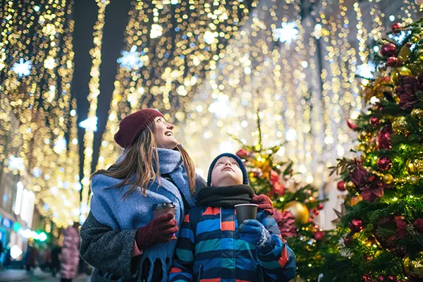 A white woman a white boy in winter coats, wooly hats and scarves, holding hot drinks and standing in front of a twinkling Christmas tree and huge blankets of lights. 