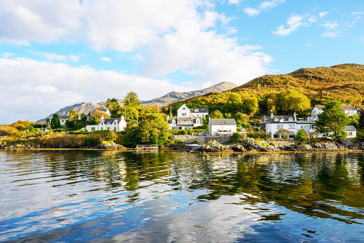 A picturesque village of white houses on the banks of a loch in Scotland, with green hills and blue sky in the background.