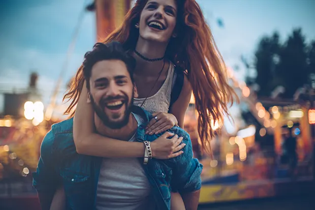 A 20-something white man with a beard giving a 20-something white woman with red hair a piggy back at a fairground at dusk.