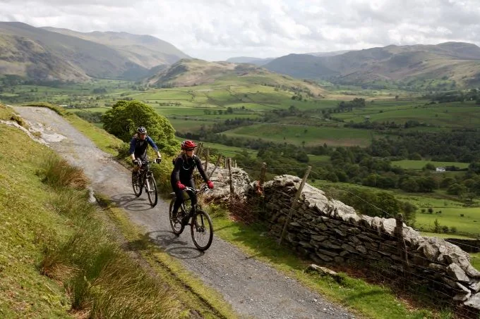 Two cyclists on their bikes making their way down a narrow pathway with green rolling hills and mountains in the background.