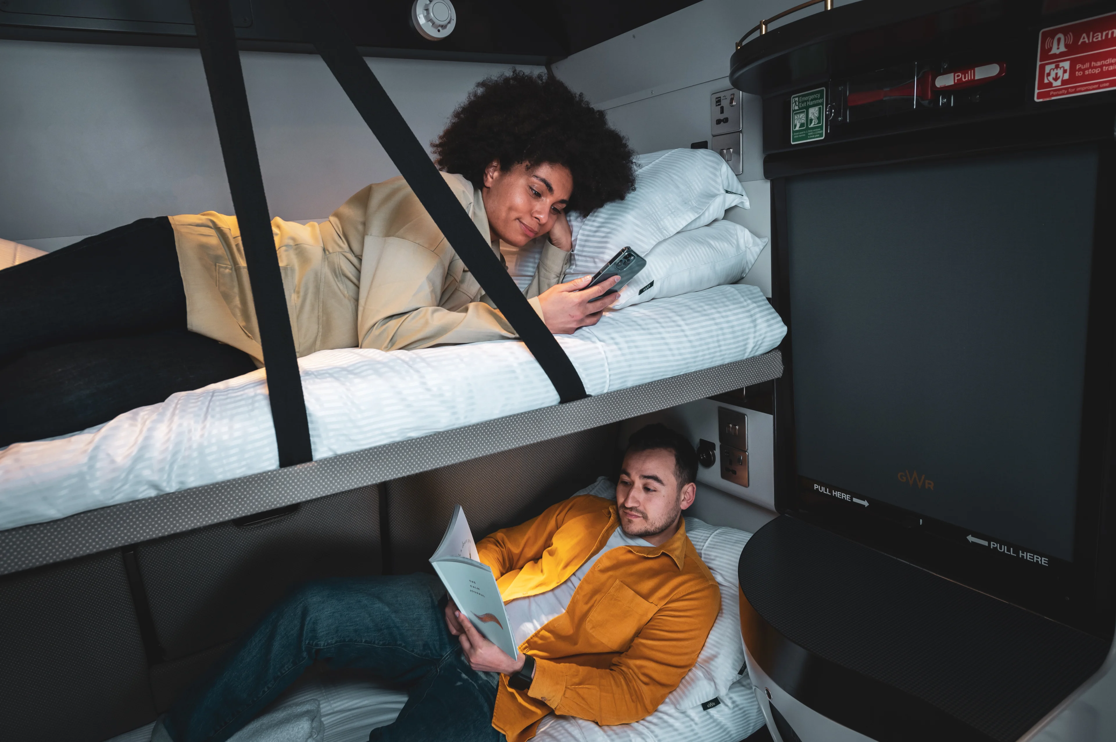 In a sleeper train carriage, a young woman lies on the top bunk looking at her phone, while a young man lies on the bottom bunk reading a book