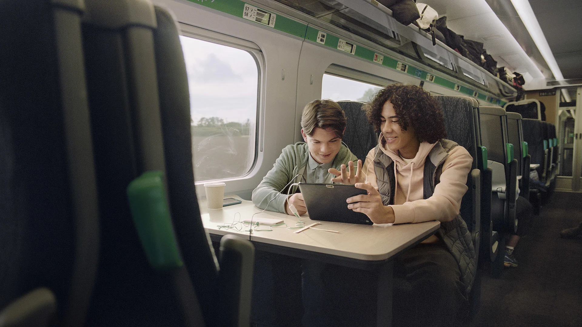 Two young men sitting at a train table, they are both smiling while looking at tablet and wearing headphones