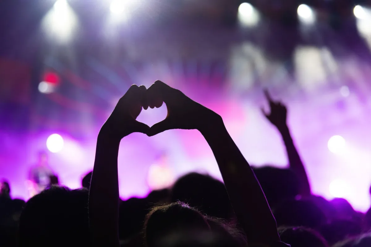 A silhouetted pair of hands forming a heart shape at a live concert, with purple and white stage lights in the background.