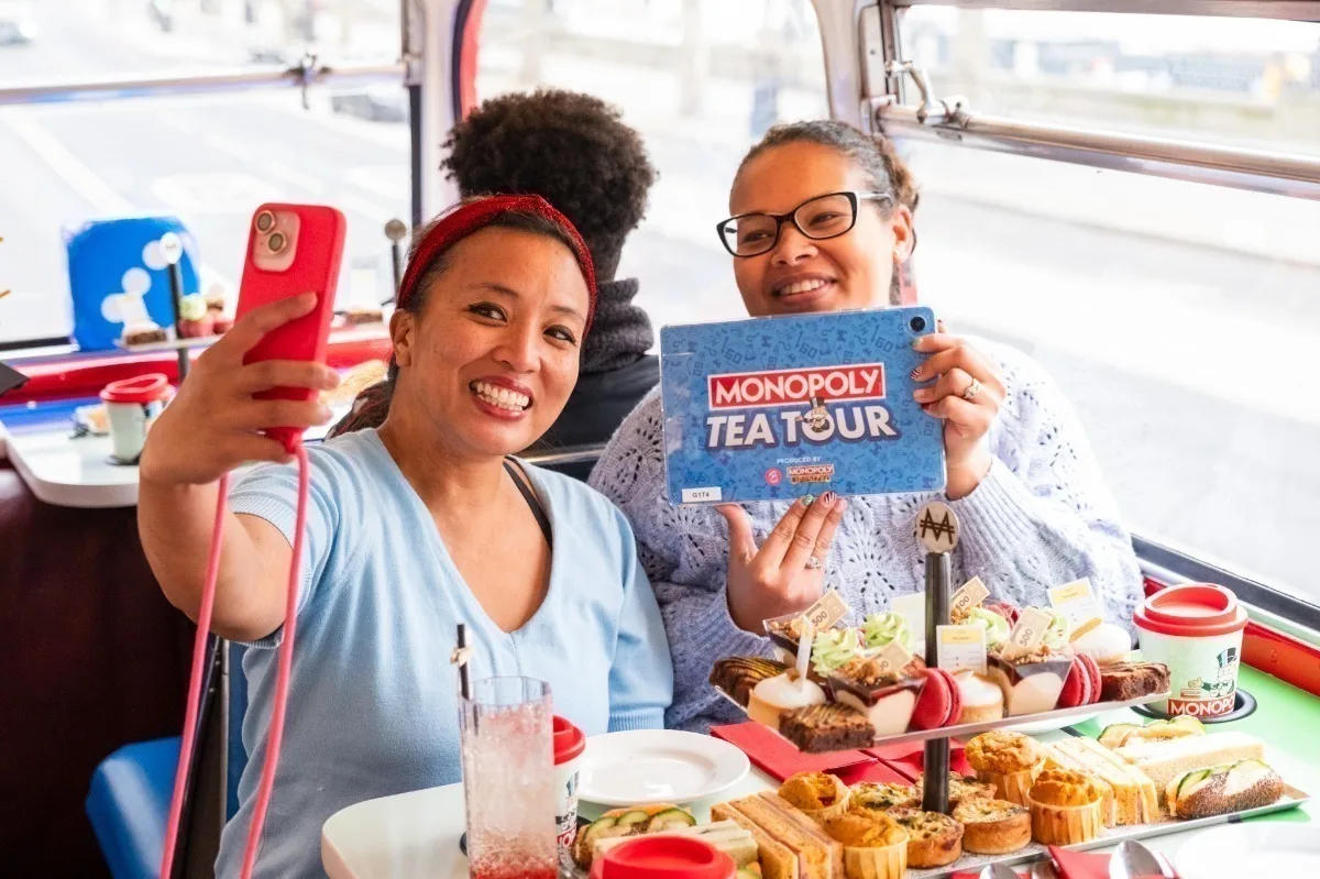 2 Black women sitting on the top deck of a double decker bus with afternoon tea on a table in front of them. One woman holds a card that says MONOPOLY TEA TOUR and the other is holding a phone to take a selfie. 