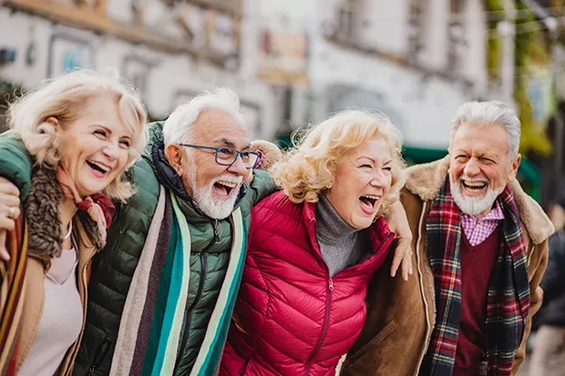 A group of 4 white senior-age friends with their arms around each other, smiling and laughing. 