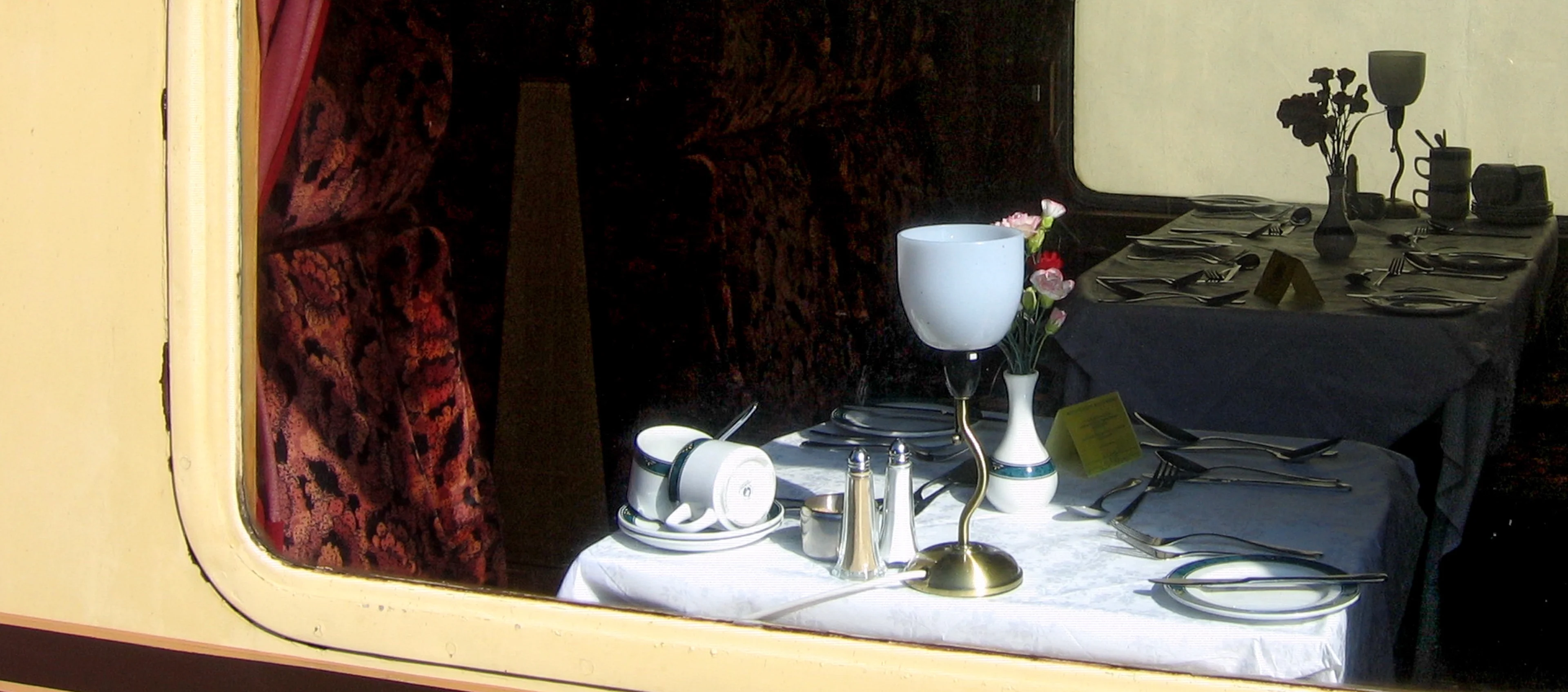 Looking into a train Pullman dining car from outside, the table is laid with a white tablecloth and crockery and cutlery, with flowers in a vase