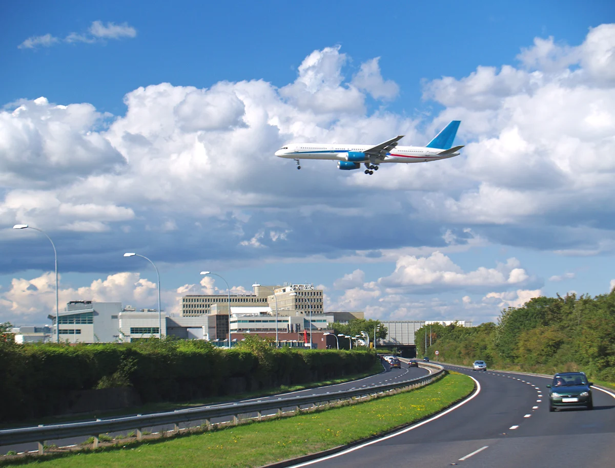 A white aeroplane in the sky above Gatwick airport, a road with trees is on one side, the terminal building is in the background. 