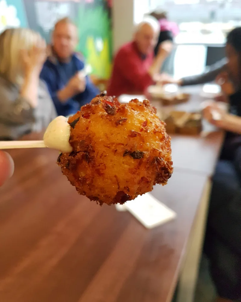 A battered and fried dumpling on a stick, with people sitting around a restaurant table out of focus in the background. 