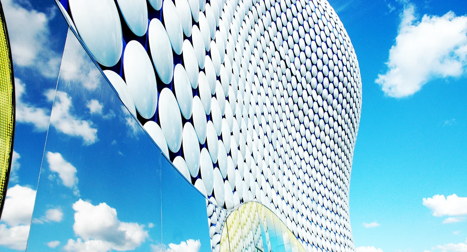A curved building covered in white circular panels, with blue sky and clouds reflected in a mirrored surface on its side.