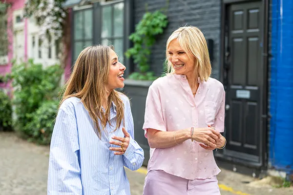 Mel Blatt, a white woman with long brown hair, and Jo Whiley, a white woman with blonde hair, walking together on a London street. 
