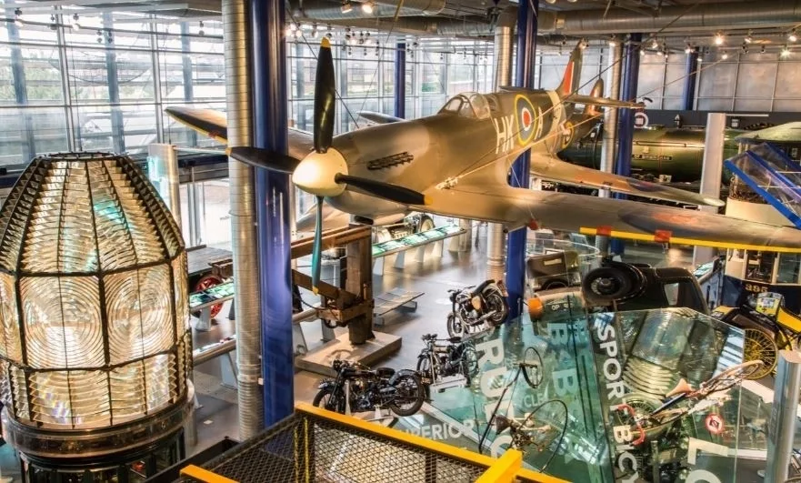 Looking across a museum gallery with an aircraft suspended from the ceiling and different exhibits, including motorbikes, on the floor. 