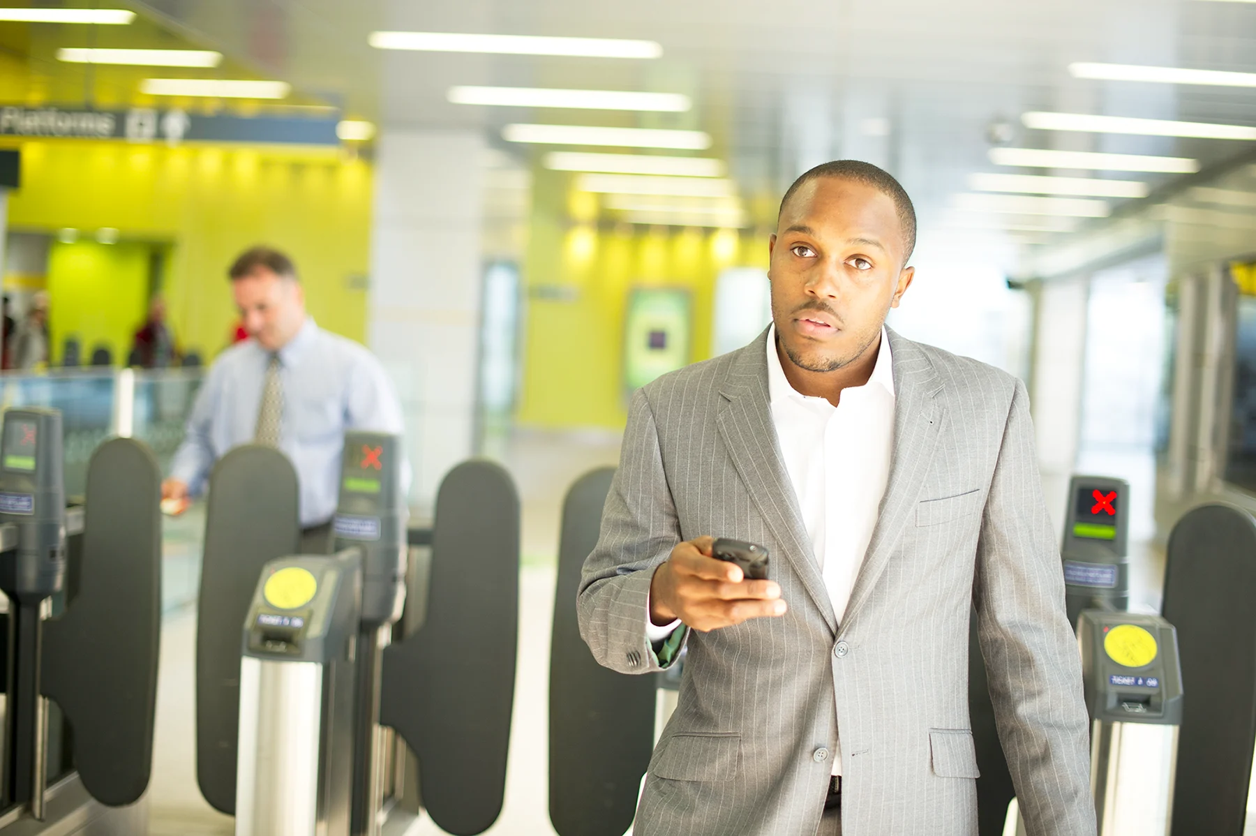 A Black man in a shirt and suit jacket holds his mobile phone by a train station ticket barrier