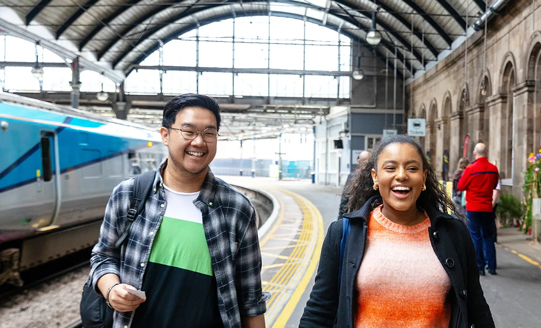 2 young people, a South Asian man and a Black woman, dressed in casual clothes and laughing and smiling as they walk along a platform at a train station. 