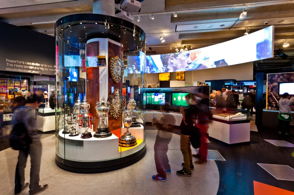Visitors to a museum looking at football trophies in a large glass cabinet. 