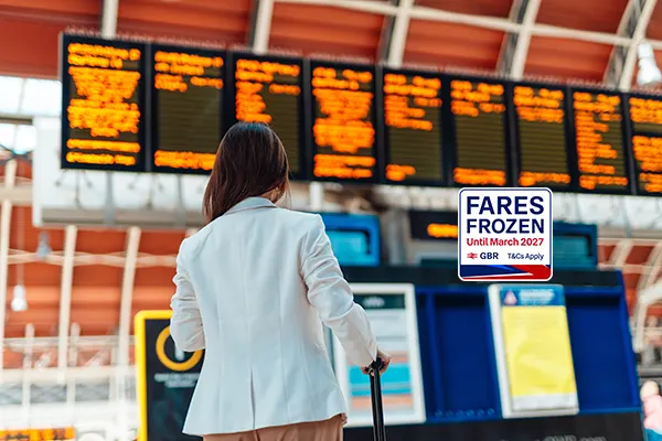 Person in white jacket looking at departure board with yellow text displays at railway station. A badge is over the image, which says FARES FROZEN UNTIL MARCH 2027.