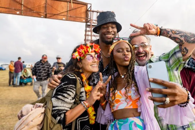 Group of friends consisting of two women, one black one white in colourful attire, and two men, one black one white, share peace signs while taking a selfie in front of the entrance to a festival.