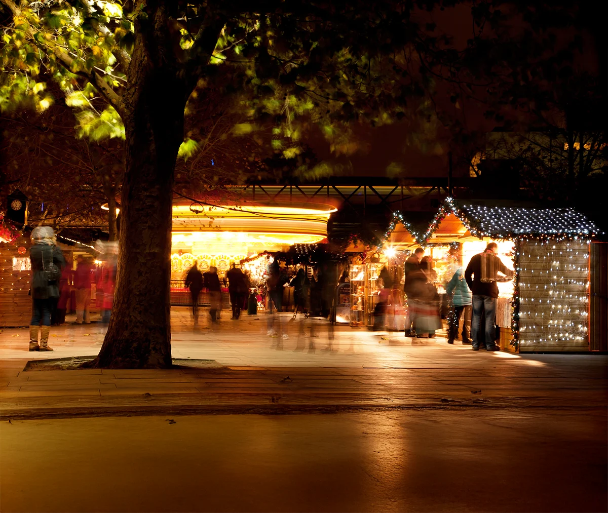 Night-time Christmas market with illuminated wooden stalls and blurred image of shoppers beneath a tree.
