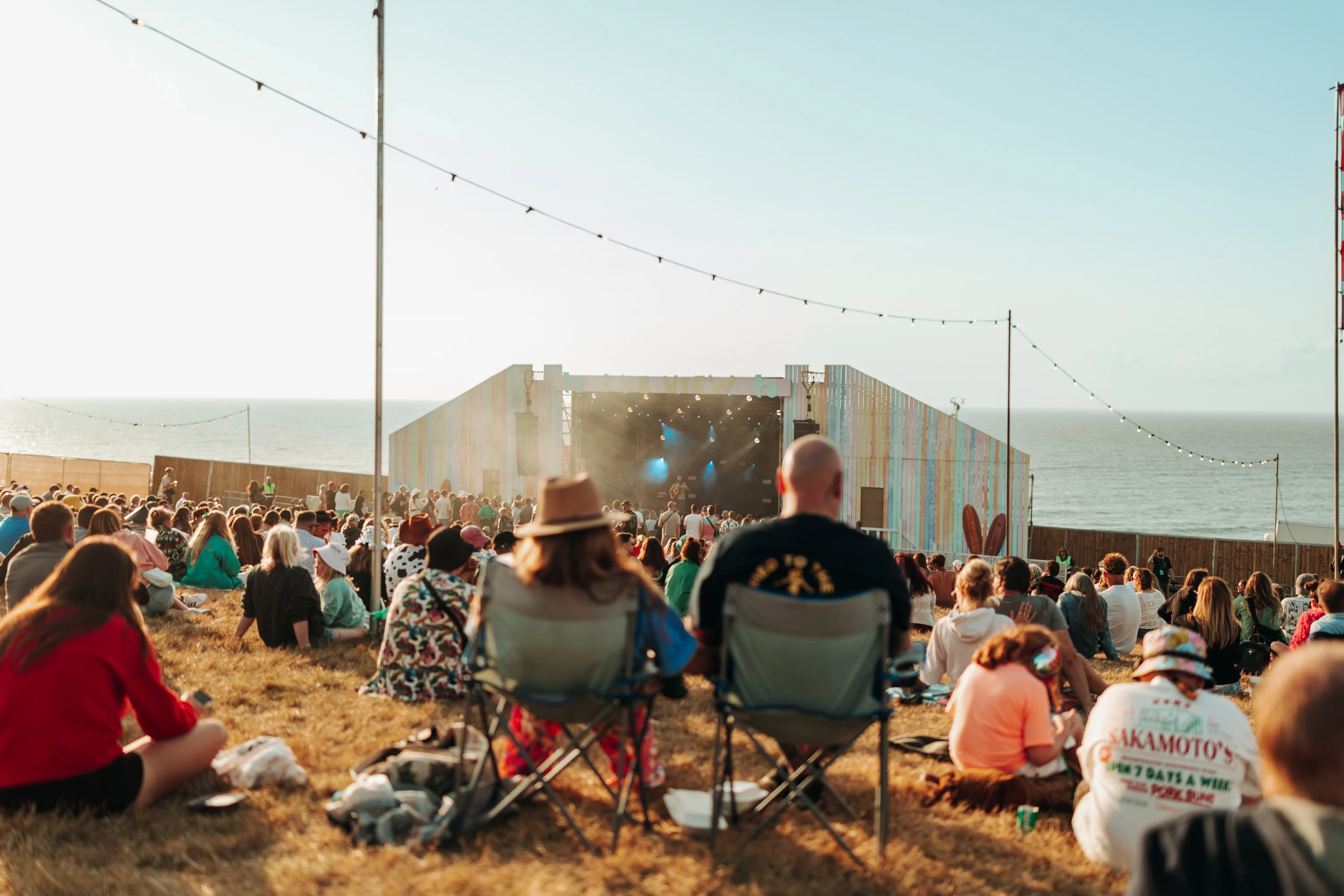 festival attendees sit amongts other revellers on a hill, facing a stage and overlooking the sea at Boardmasters festival