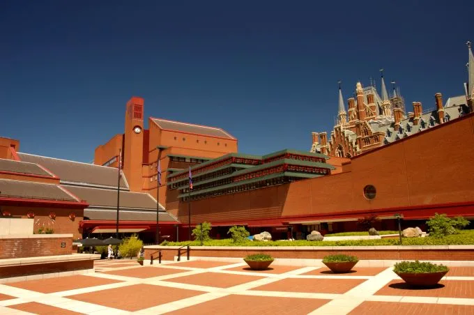 Building with terra cotta colour bricks sit beneath a blue sky. Square large tiles line the foreground with cream lining. Building with dramatic turrets sits in the background behind the library building.