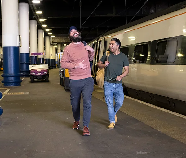 2 men (comedians Joe Wilkinson and Nick Mohammed) laughing and walking along a train platform, with a white passenger train behind them.