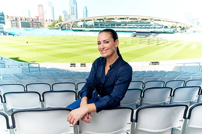 Kirsty Gallacher, a white woman with long dark hair pulled back in a ponytail, sitting in an empty sports stadium, she is smiling and looking at the camera.