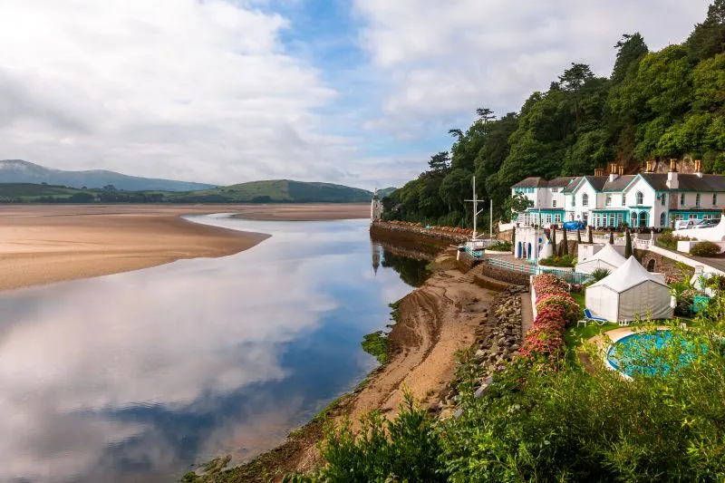 A small village scene of white boroque buildings sit to the right, overlooking the sea in the middle of the image, and a sandy beach parallel to to the buildings, is on the left of the image.
