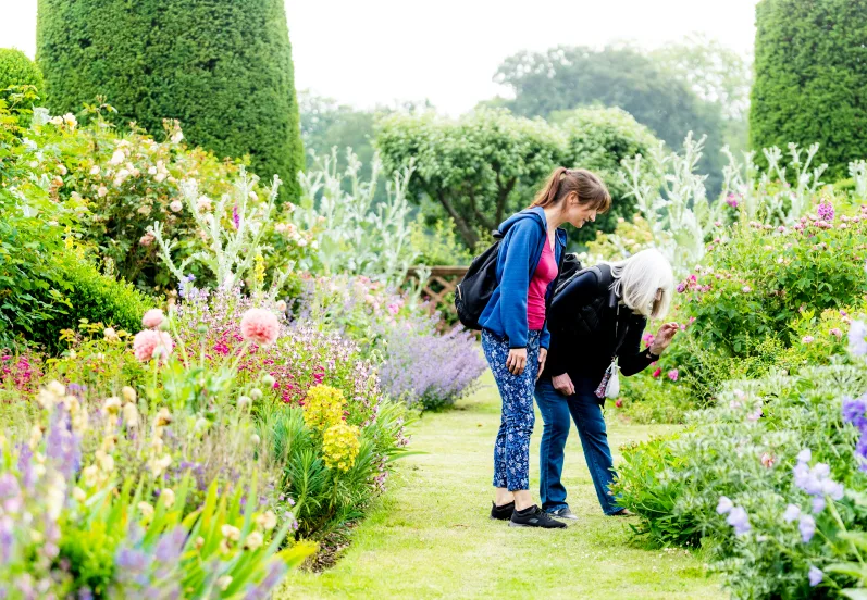 2 white women standing in a well-maintained formal garden with topiary and colourful flower beds. 1 woman is bending down to take a closer look at a flower. 