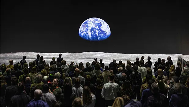A large group of people standing in front of a huge projection showing a black sky and the Earth from space. 