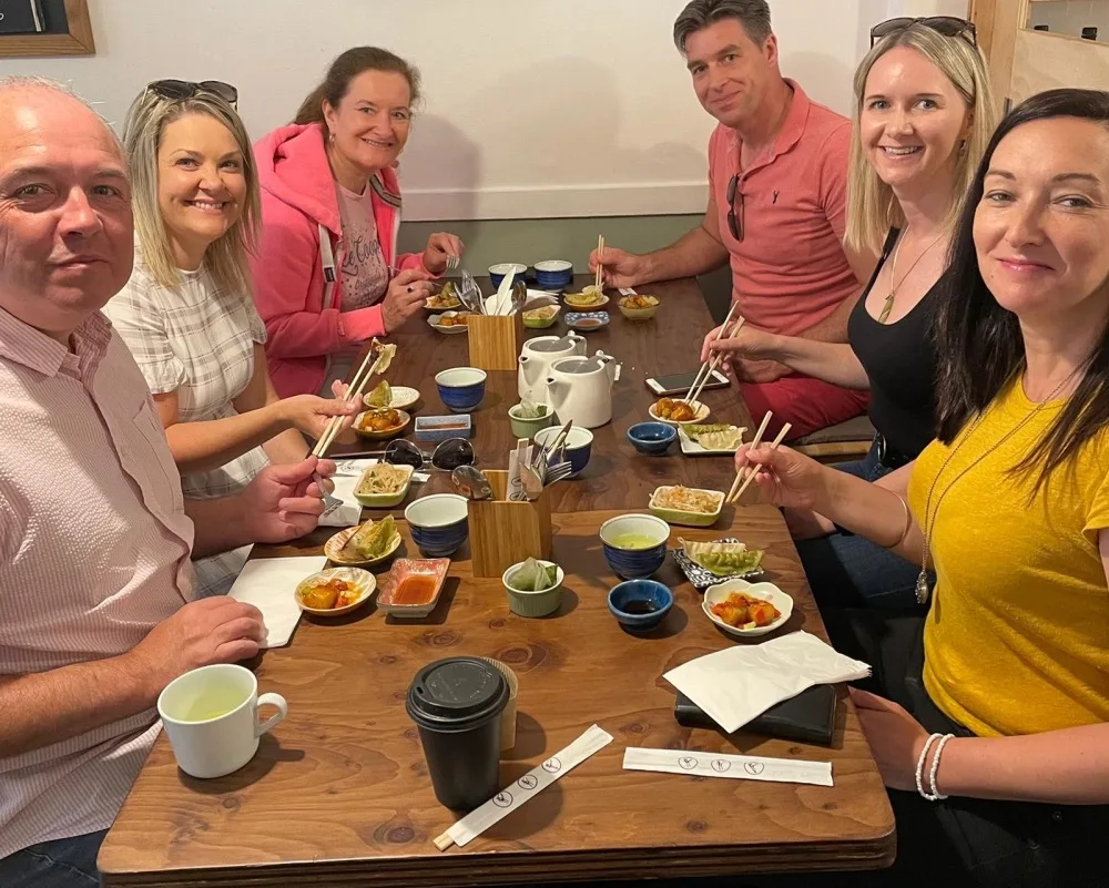 6 white people sitting at a wooden restaurant table and holding chopsticks. 
