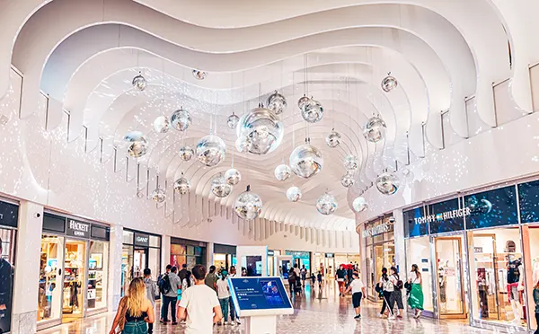 The inside of a shopping mall with a high, with a wavy ceiling design that has multiple large silver balls hanging from it. 