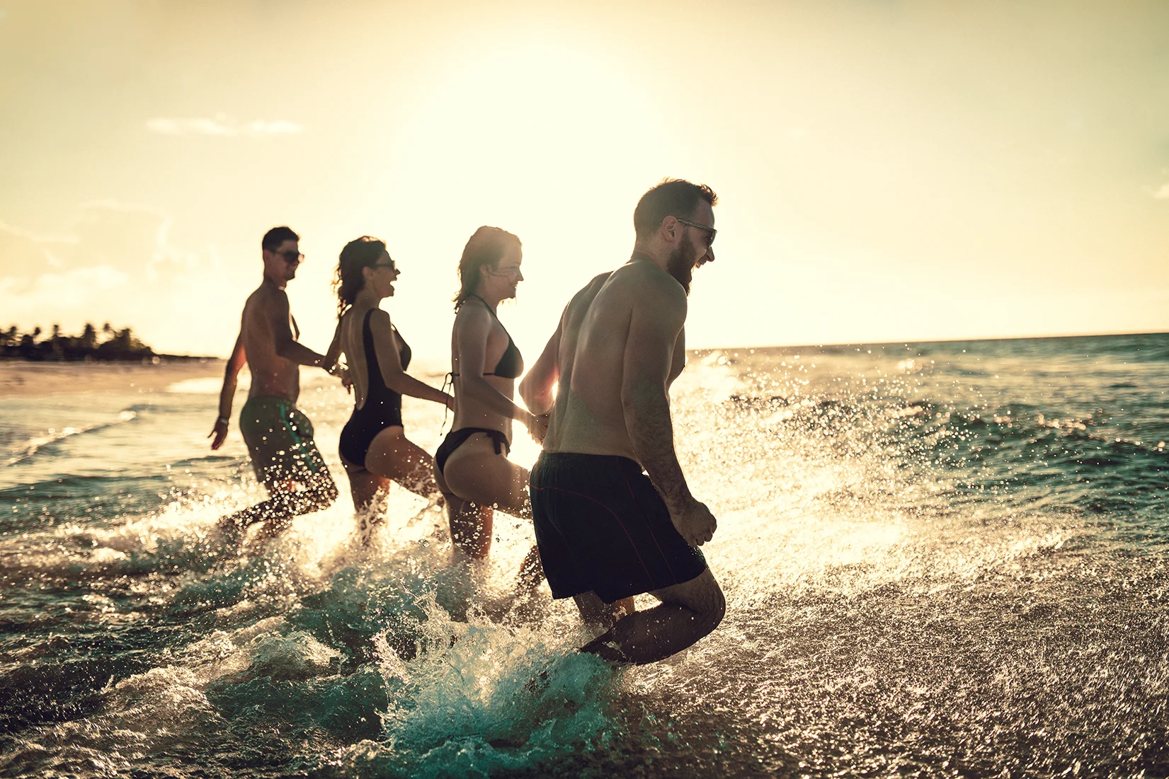 Four friends holding hands and wearing swimwear walk into the sea on a sunny day
