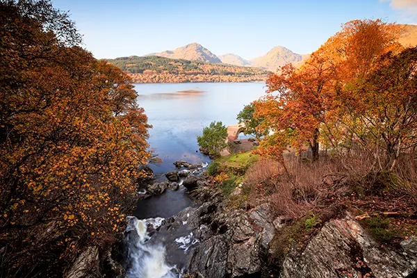 Looking across rugged river banks with a waterfall and autumn trees to a large loch and mountains in the distance. 