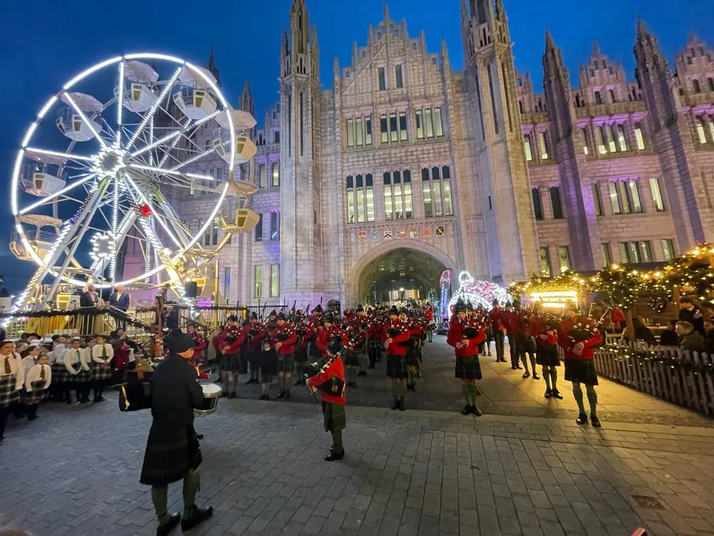 A band of Scottish pipers in front of a cathedral illuminated for Christmas.